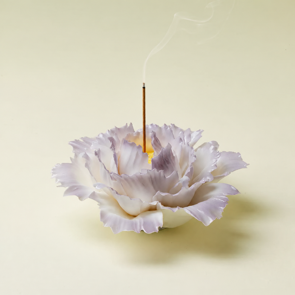 A white and pale lavender flower-shaped incense holder with a single burning incense stick rising into the air, set against a solid pale cream background.