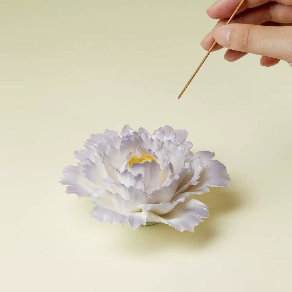 A pale lavender and white peony flower with a yellow center, shown on a light beige background with a hand holding a wooden stick above it.