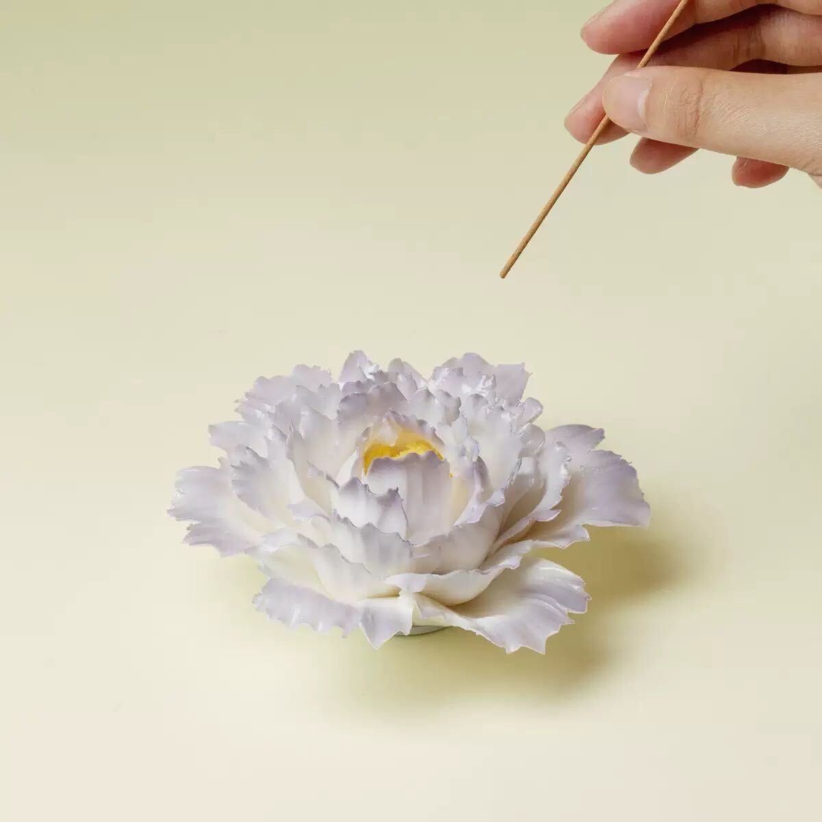 A pale lavender and white peony flower with a yellow center, shown on a light beige background with a hand holding a wooden stick above it.